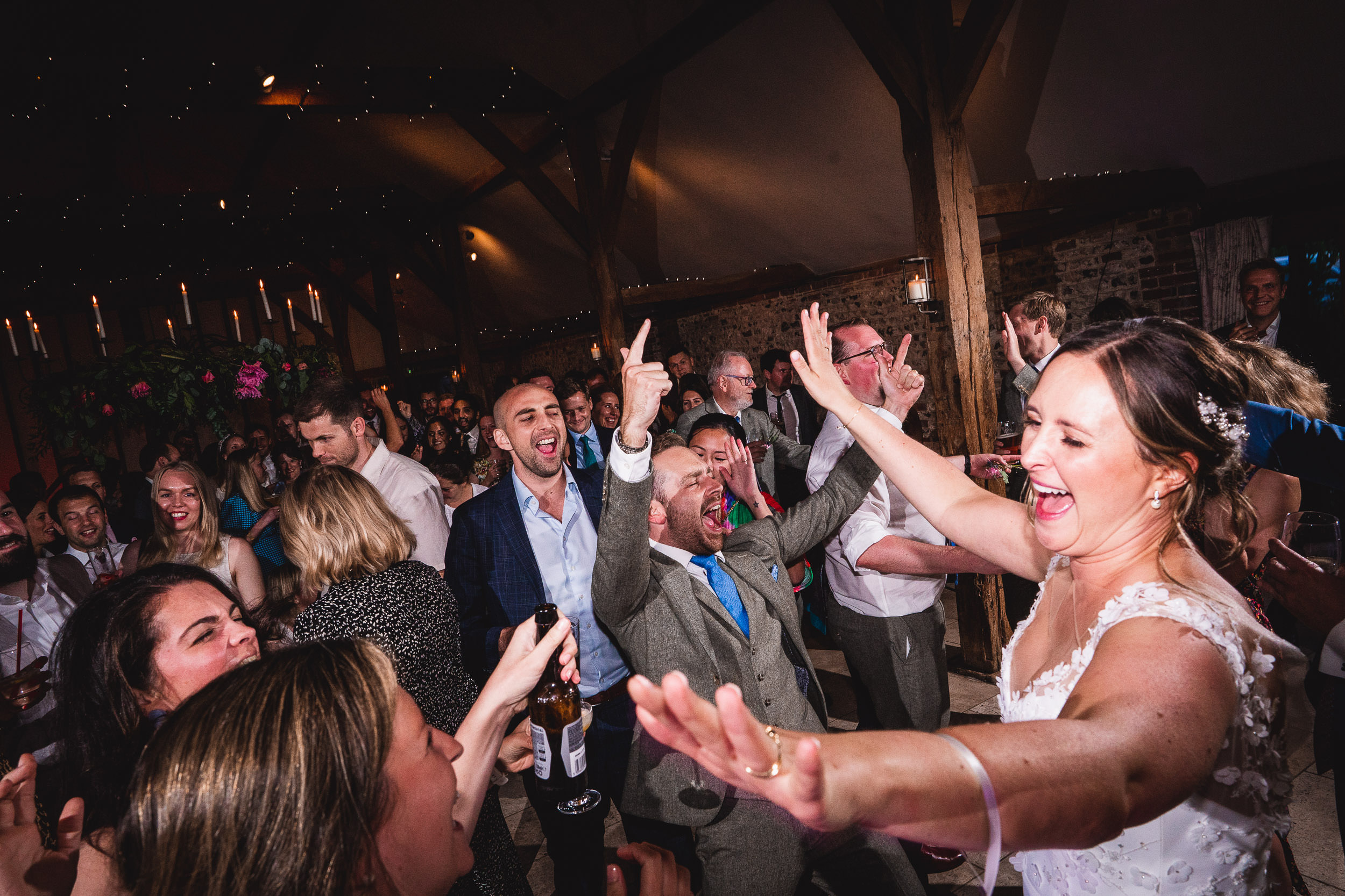 A joyous bride and guests dance energetically at a lively indoor wedding reception, surrounded by wooden beams and festive lights.