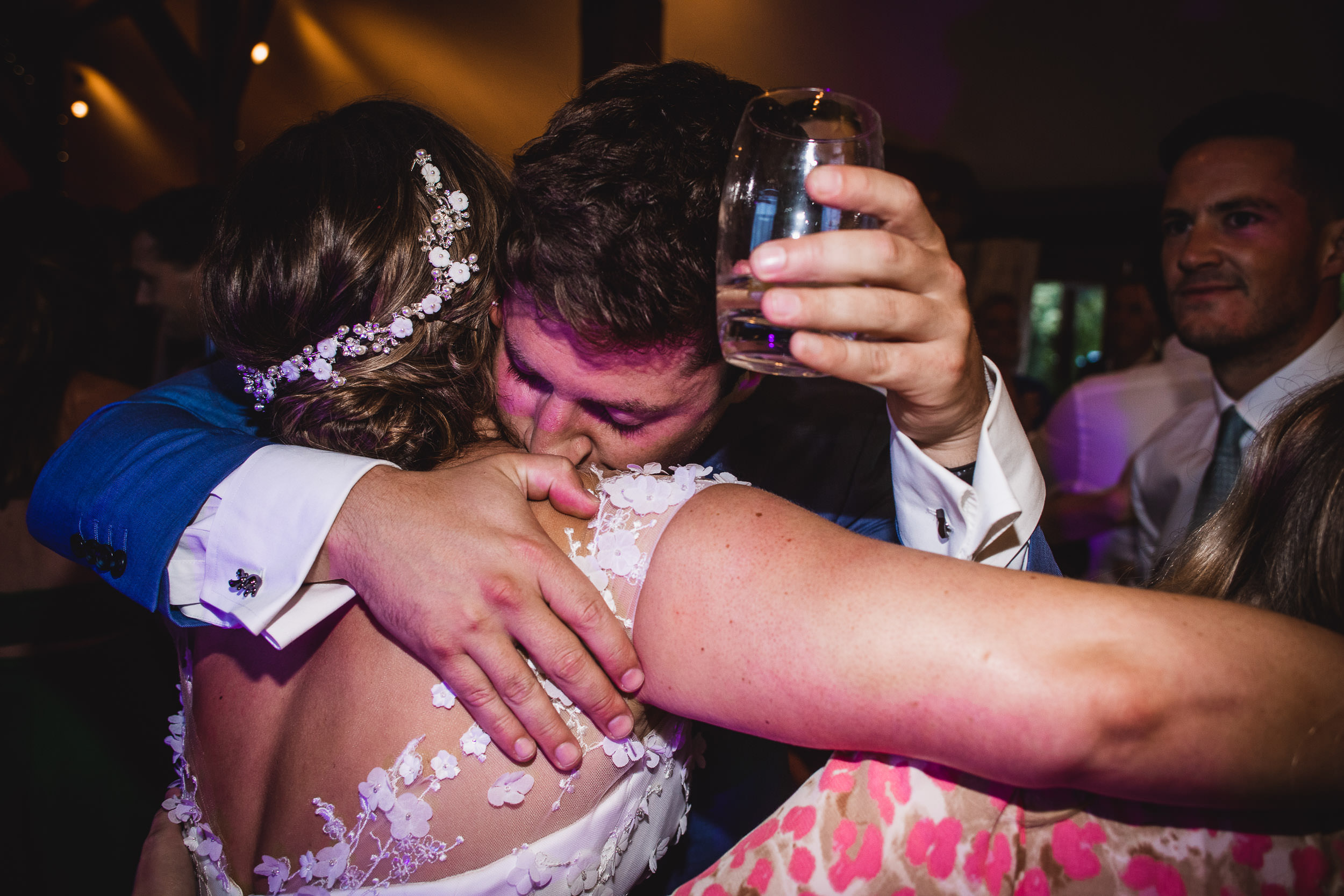 A person in a blue suit hugs a person wearing a white floral dress while holding a glass of water. Another person in a floral outfit is partially visible.