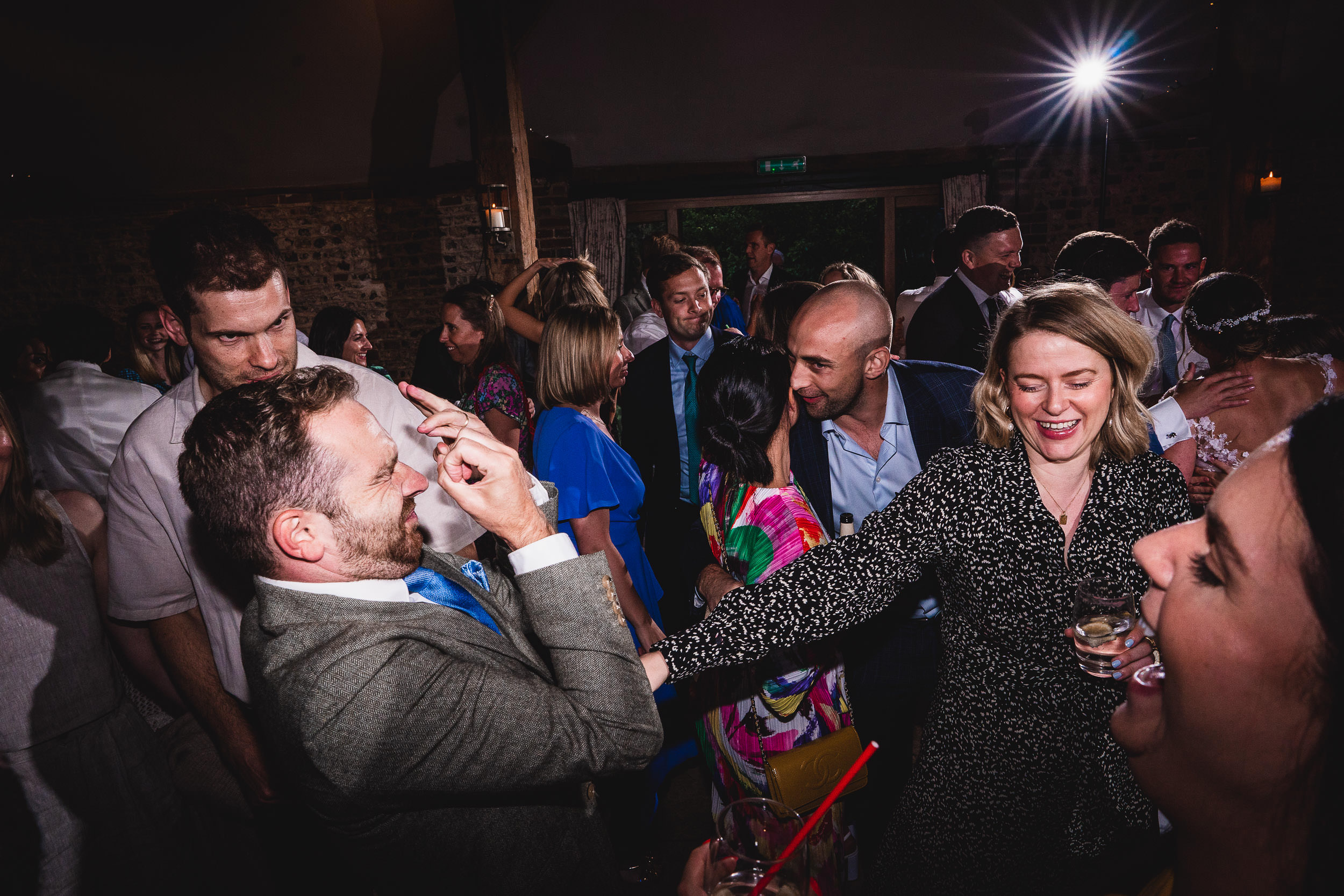 A lively indoor party with people dancing and laughing, some holding drinks. A man in the foreground playfully gestures towards a woman smiling with a glass. Bright lighting accentuates the scene.