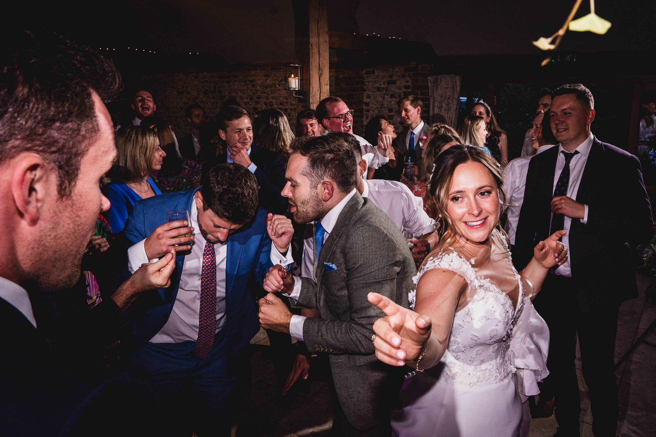 A bride in a white dress and guests dance energetically at a wedding reception, with a man in a blue suit nearby.