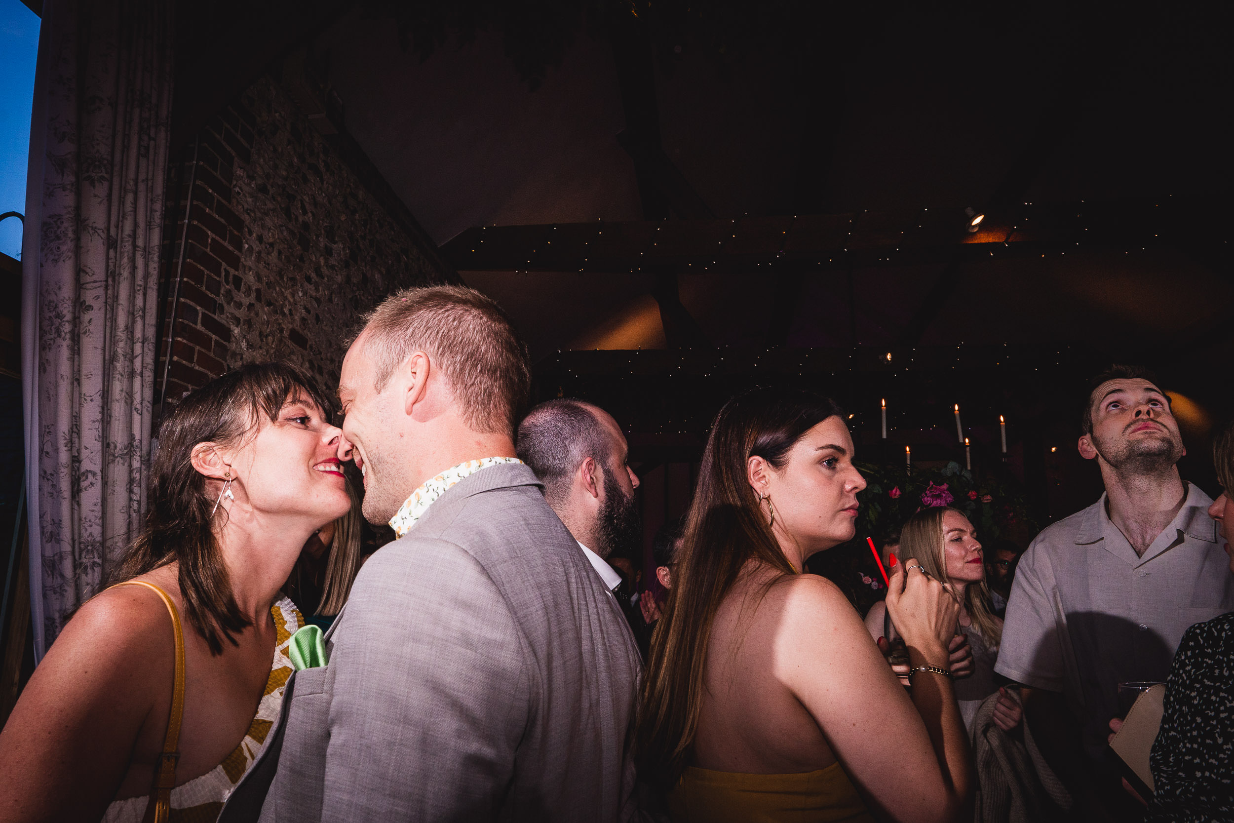 A couple leans in for a kiss at a dimly lit social gathering, surrounded by other people.