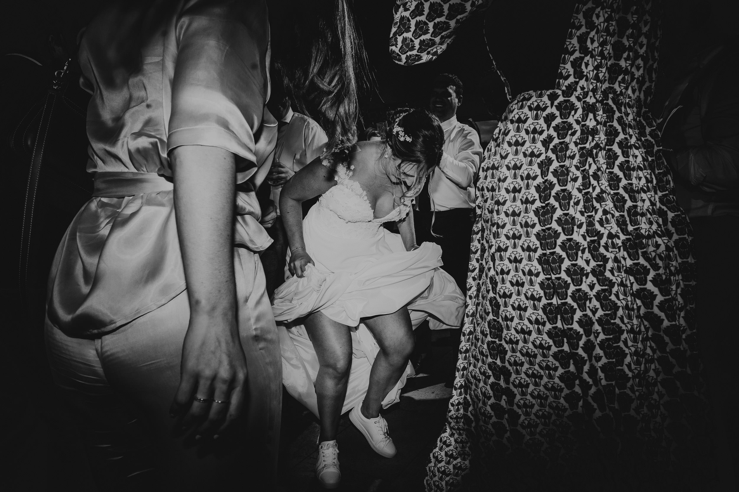 A bride energetically dances in a lively crowd, wearing white sneakers with her wedding dress. The scene is captured in black and white.