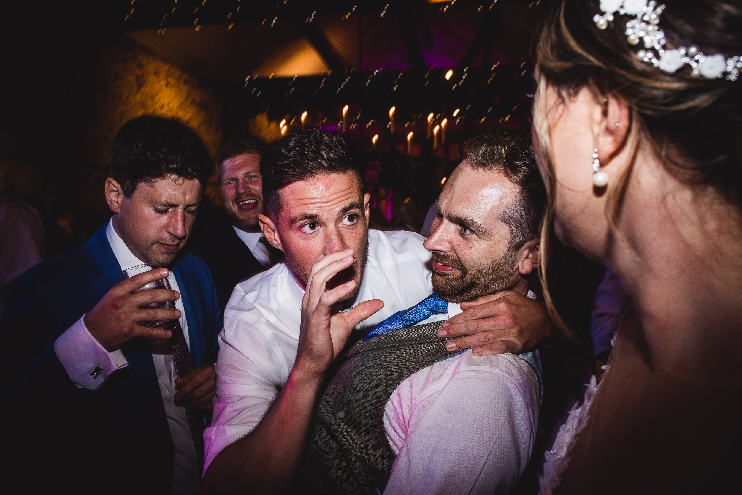 A group of people in formal attire are gathered at an event, with one person appearing to whisper to another. The background is dimly lit with string lights.