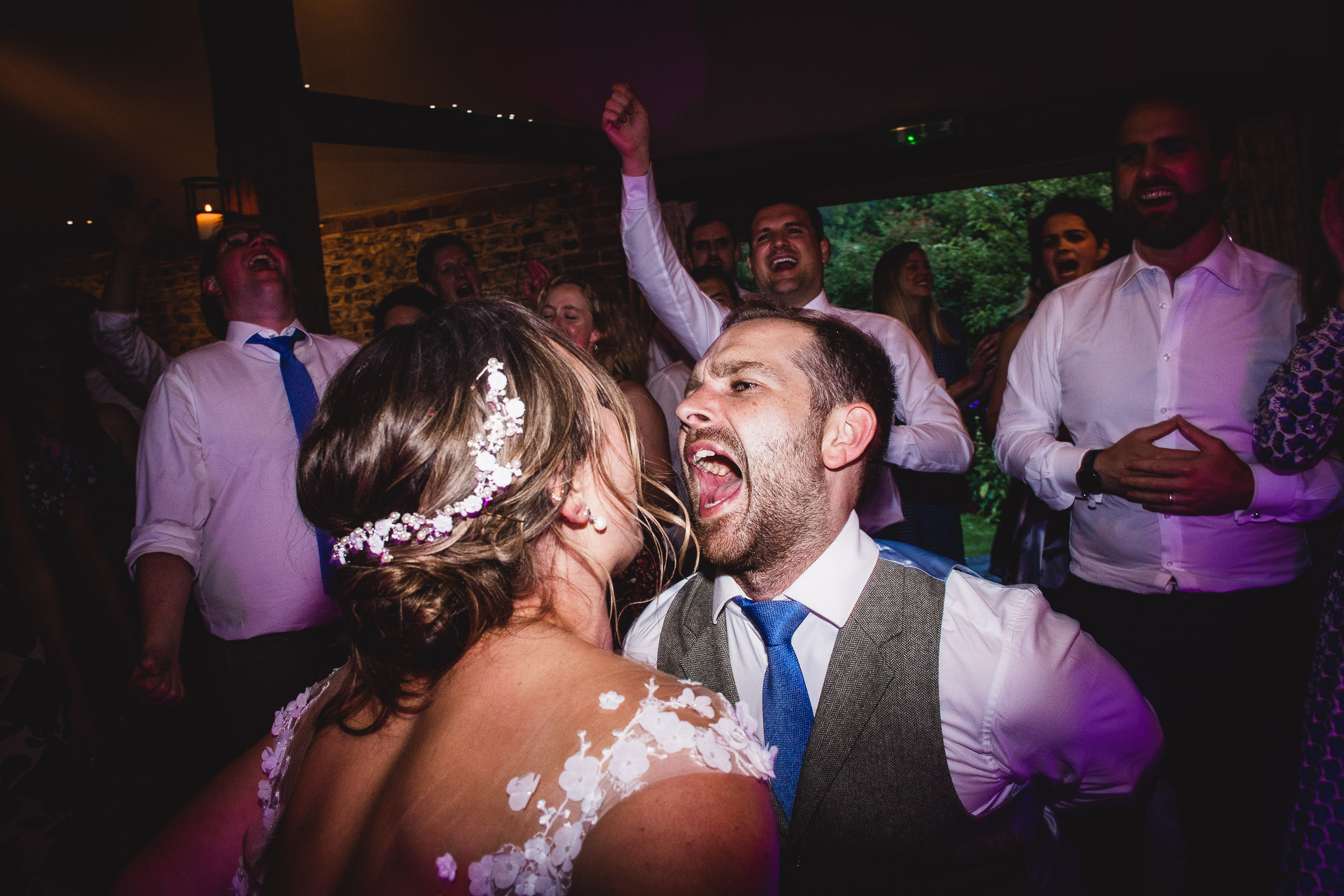 A joyous group celebrating, with a man in a vest enthusiastically singing to a woman in a floral dress. Other people in the background are also cheering and singing.