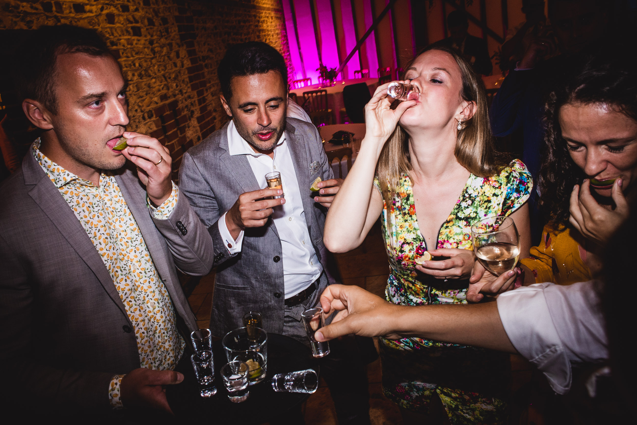 Five people in a lively setting, some holding drinks and others taking shots, with vibrant lighting and a brick wall in the background.