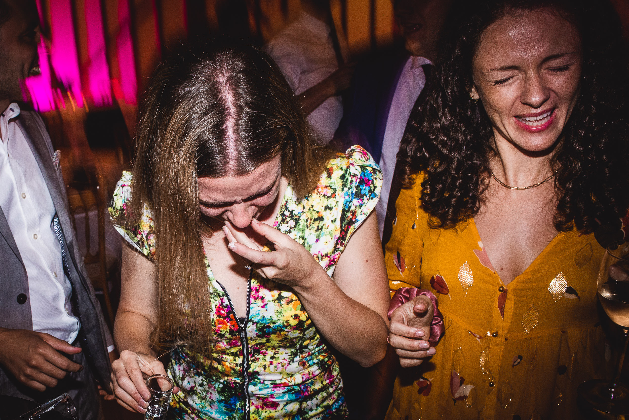 Two women laughing and holding drinks at a lively party, with vibrant lighting in the background.