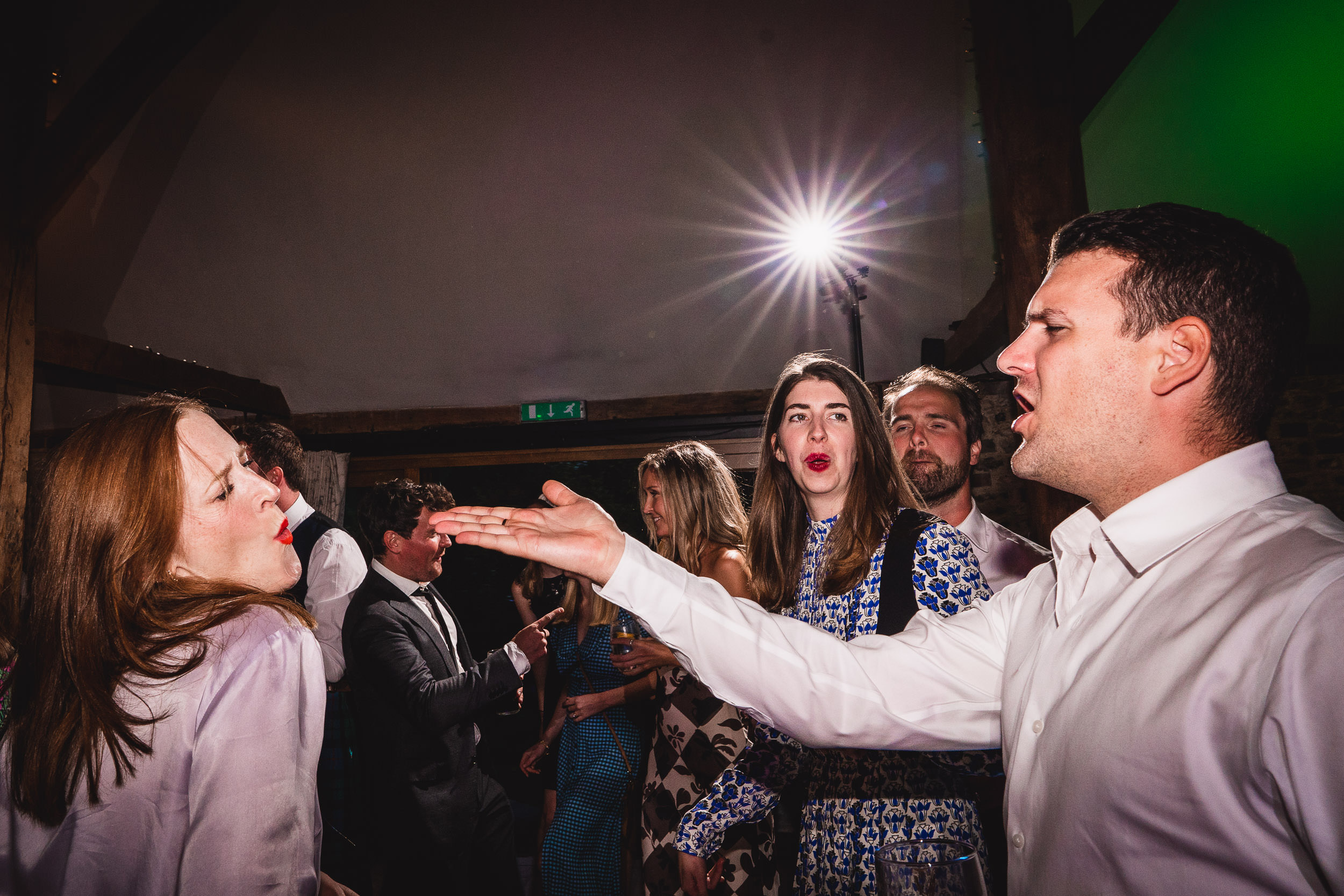 A group of people at a party are engaged in lively conversation and singing, wearing formal attire, with a bright spotlight in the background.