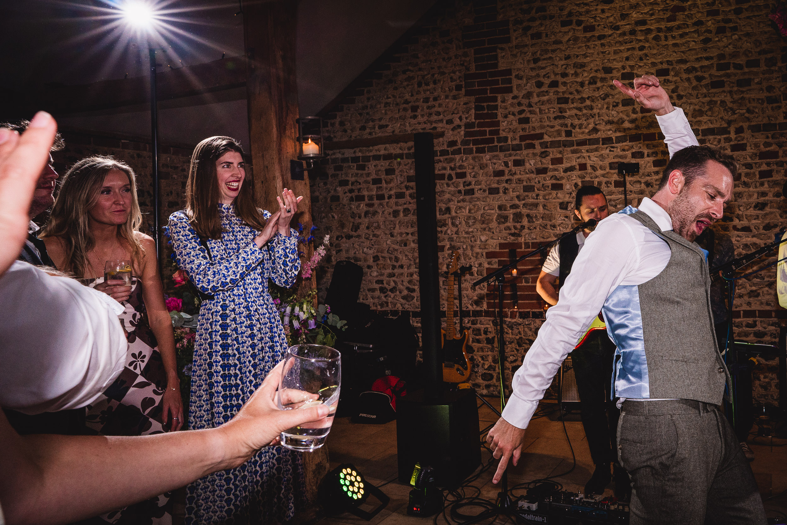 A man in a vest dances energetically at a party while others watch and clap. A woman in a blue dress is clapping. A person in the foreground holds a drink.