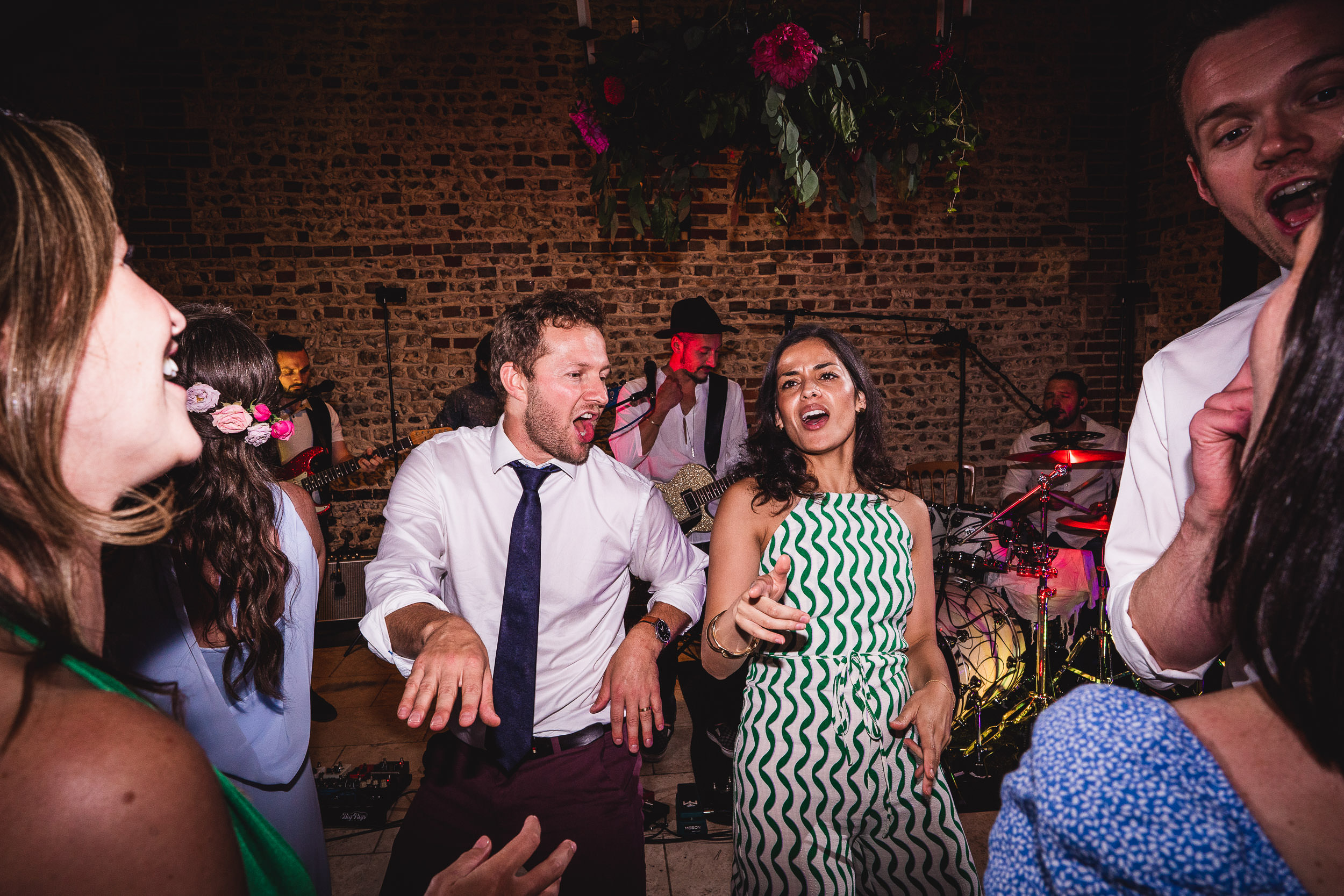 People are dancing energetically together at a lively indoor event with a band playing in the background. The setting has brick walls and hanging greenery.