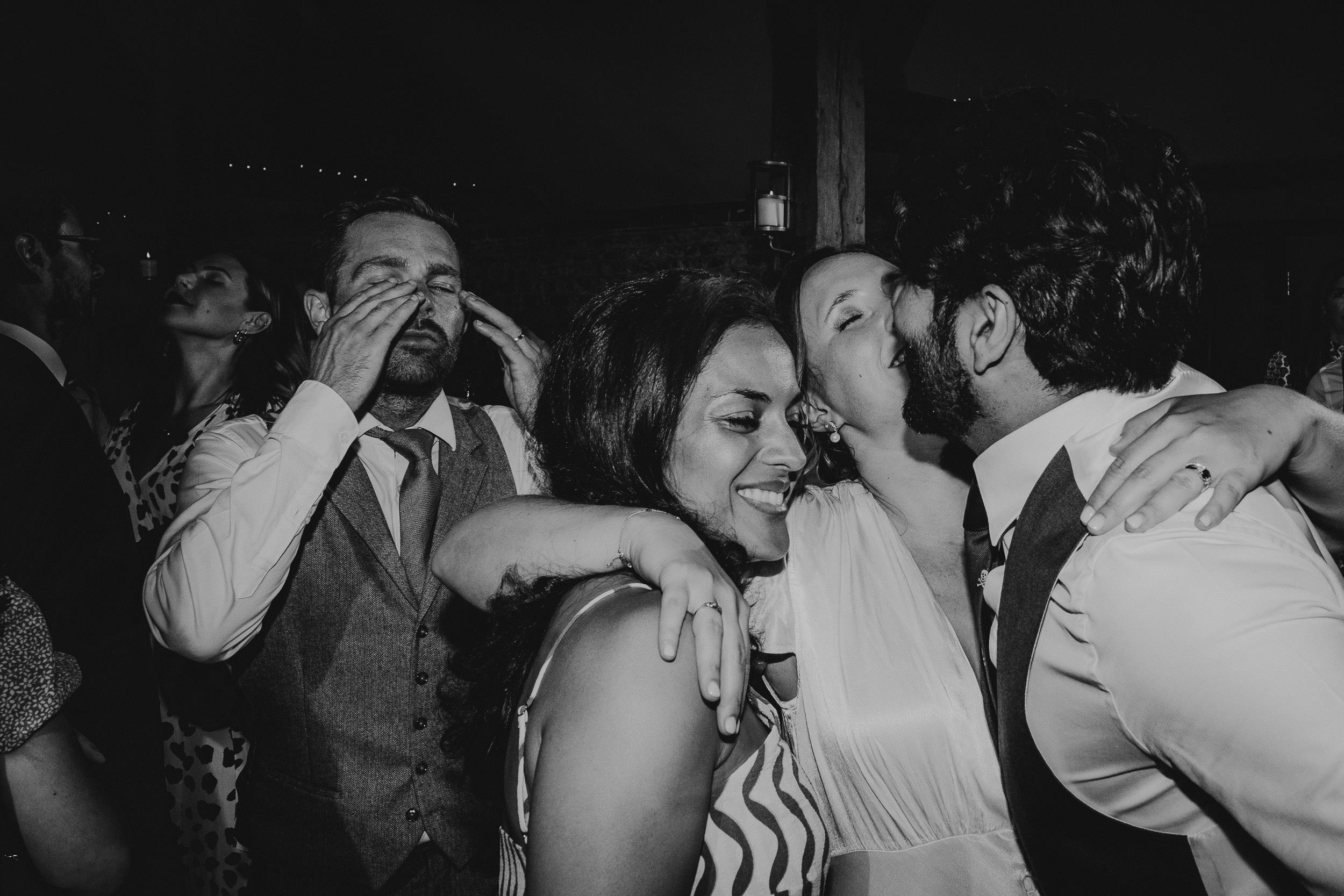 A group of people are dancing closely together at a lively event. A man and woman kiss in the foreground, while others laugh and enjoy themselves in the dimly lit setting.