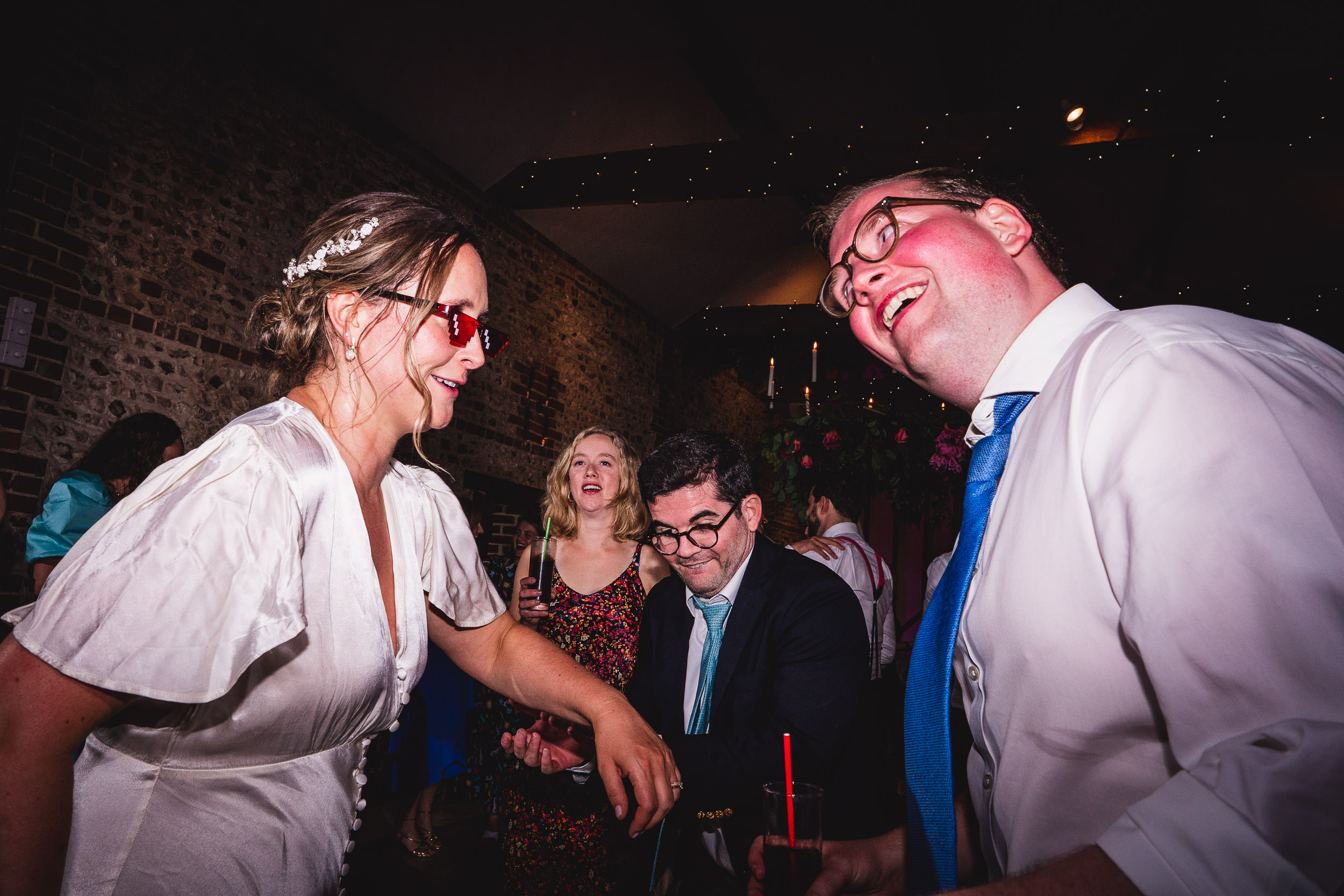 A group of people in formal attire are dancing and laughing in a dimly lit venue. Some are holding drinks, and the background includes stone walls and decorative lights.