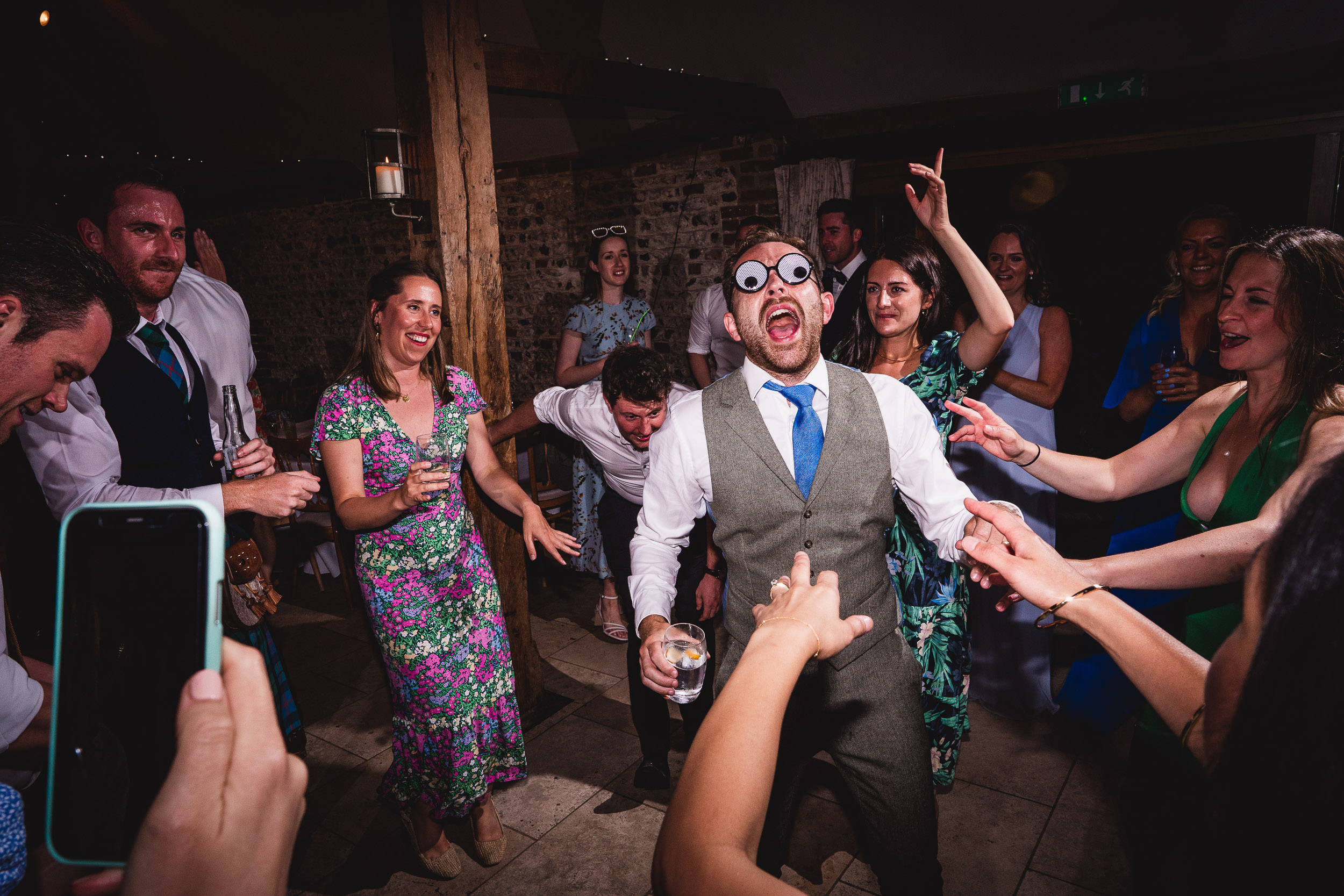 A group of people in formal attire are dancing energetically at a party. One man in the center is wearing novelty glasses, holding a drink, and surrounded by others joyfully reaching out.