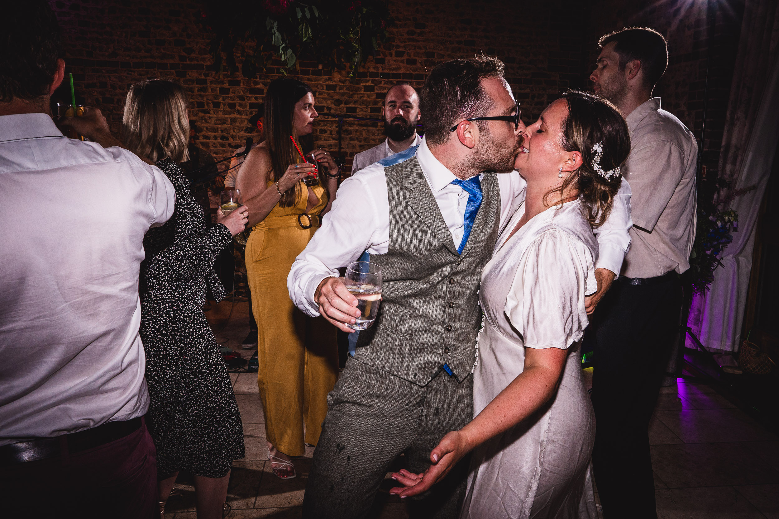 A couple kisses on a dance floor surrounded by people in formal attire. The man holds a drink in one hand. The background shows a brick wall and soft lighting.